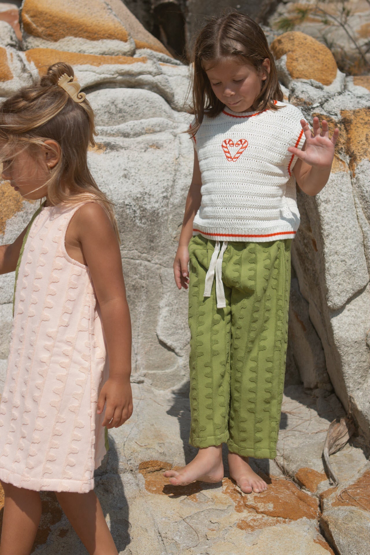 Child wearing a white knit vest with red candy canes and green towelling pants standing on a stone surface.
