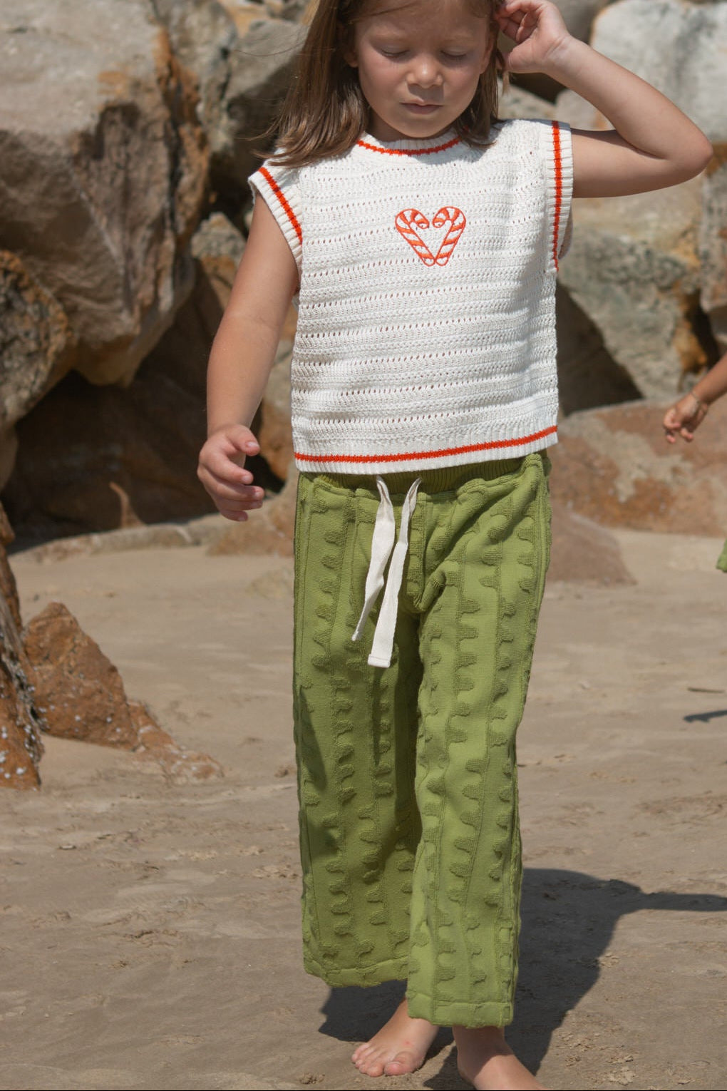 Child wearing a white knit vest with red candy canes and green towelling pants standing on a stone surface.