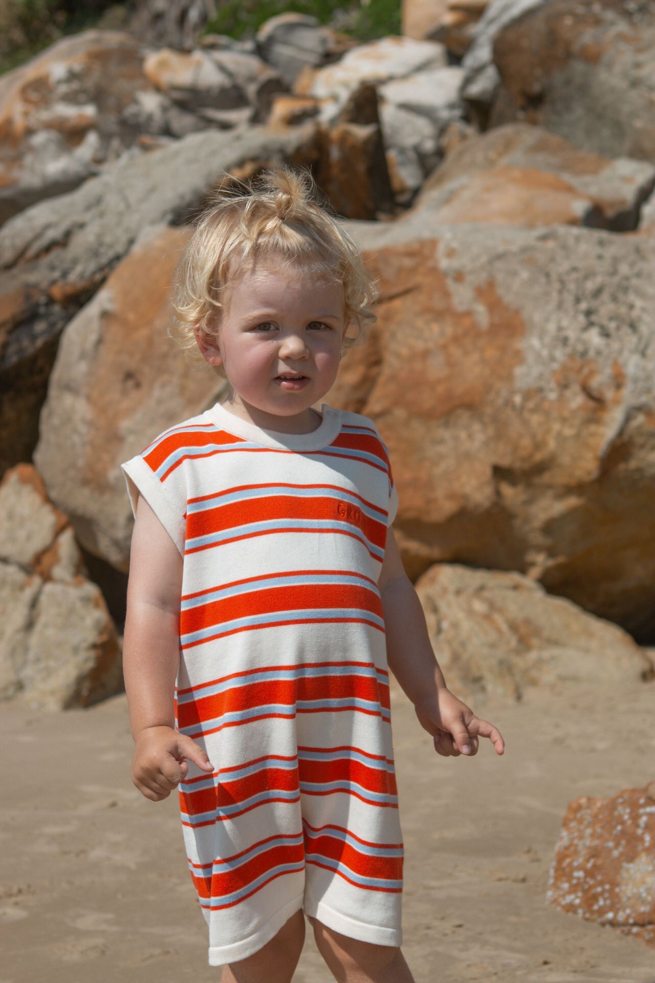 Child wearing a red and white striped romper standing on a rocky beach.