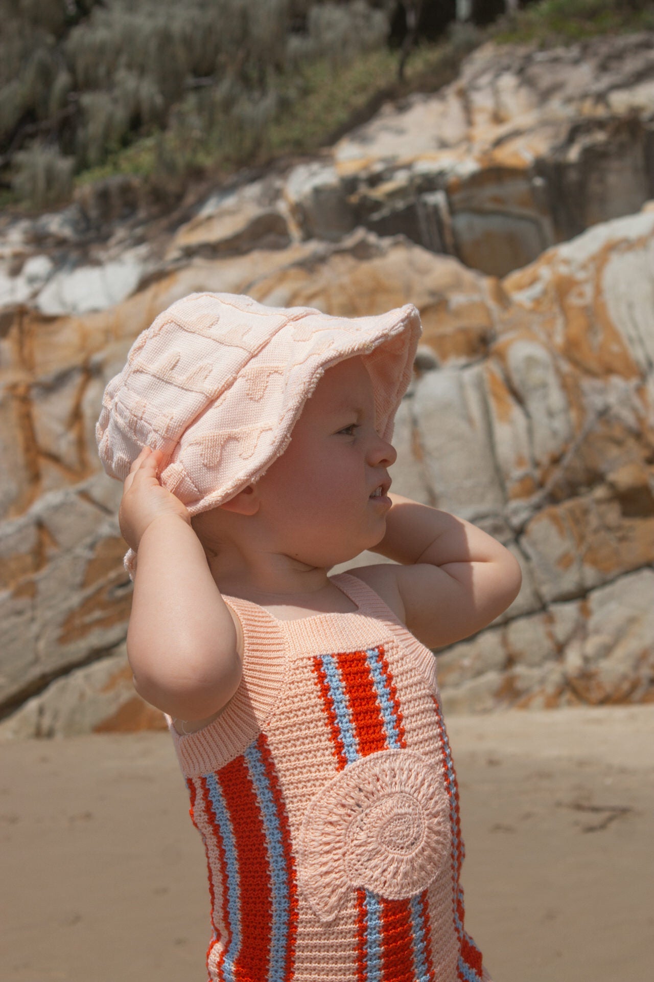 Child wearing a pink sun hat and striped singletsuit on a rocky beach.