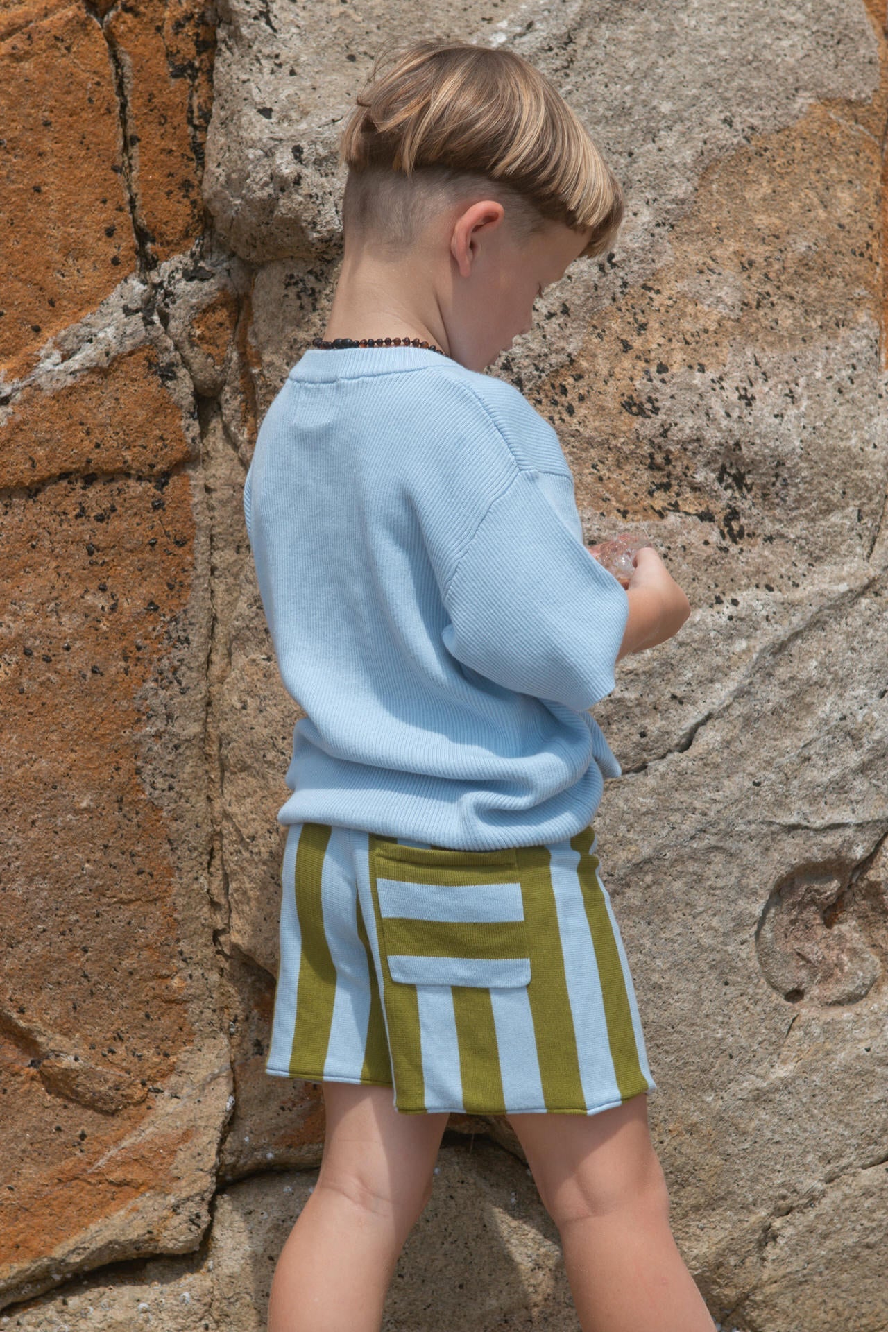 Child wearing a light blue shirt and striped shorts against a stone wall.